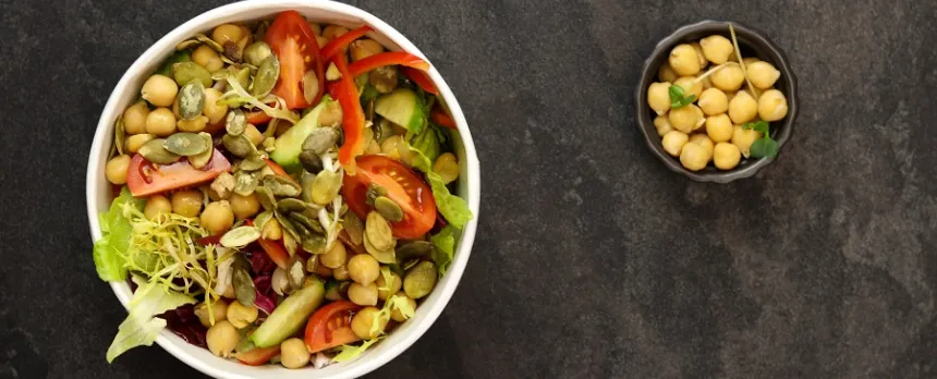 A large white bowl with chickpeas and other Mediterranean vegetables is next to a small bowl of chickpeas, both on a black background.